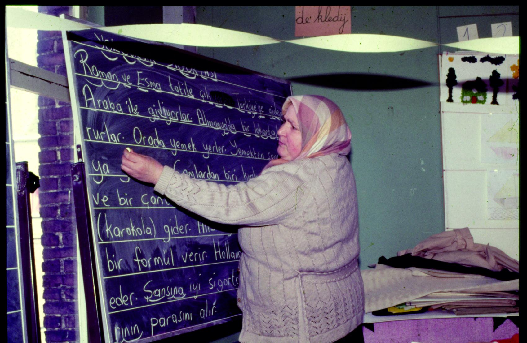 Foto van een vrouw met een hoofddoek die Turks taallessen geeft op een krijtbord.